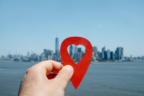 the hand of a man holding a red marker with a heart-shaped hole pointing the Financial District, in Manhattan, New York, United States, as is seen since the bay, in the foreground