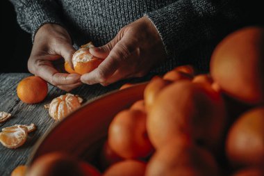 Gri kazaklı beyaz bir adam gri bir ahşap masada mandalina soyar.