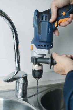 Mavi tulumlu beyaz bir adam, metal mutfak lavabosunda matkapla delik açıyor.