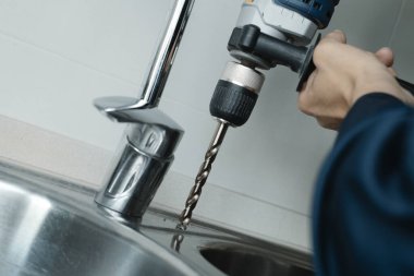 Kafkasyalı bir adamın yakın çekimi, mavi tulumlu, metal bir mutfak lavabosunda matkapla delik açıyor.