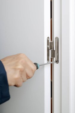 Kafkasyalı bir adamın yakın çekimi, mavi tulum giyiyor, tornavida kullanarak kapının menteşesini söküyor ya da düzüyor.