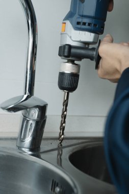 Kafkasyalı bir adamın yakın çekimi, mavi tulumlu, metal mutfak lavabosunda delik açmak için matkap kullanıyor.