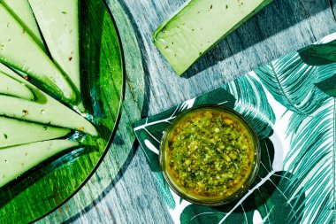 Yeşil cam bir tabağın yüksek açılı manzarası ve yanında pesto peyniri dilimleri olan cam bir kâse ve üzerinde pesto sosu olan gri bir ahşap masa.