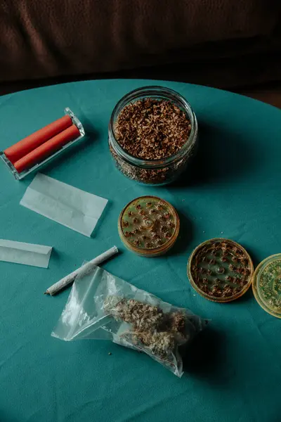 stock image some cannabis buds in a plastic bag on a table next to a used herb grinder, a spliff or joint, some rolling paper sheets and a jar with some rolling tobacco