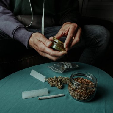 closeup of a man about to shred a cannabis bud with a used grinder, sitting on a sofa, next to a table with some rolling tobacco and rolling paper clipart