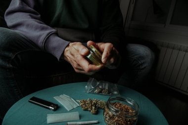 Kanepede oturan bir adam kullanılmış bir öğütücüyle esrar tomurcuğunu öğütüyor. Yanında rulo tütün ve kağıt olan bir masa var.