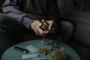 a man, sitting on a sofa, shredding a cannabis bud with a used grinder, next to a table with rolling tobacco and paper clipart
