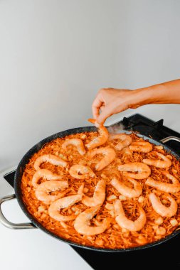 a man ads some shrimps to a spanish fideua, a noodle paella, cooked in a paella pan, in the stove of a domestic kitchen clipart