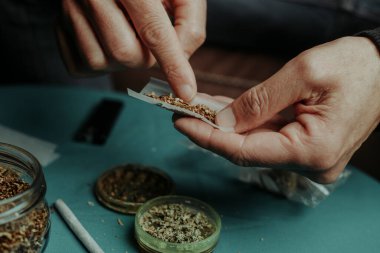 Tütünü öğütücüde öğütülmüş kenevirle karıştırmadan önce sarma kağıdına biraz tütün koyan bir adamın yakın plan fotoğrafı.