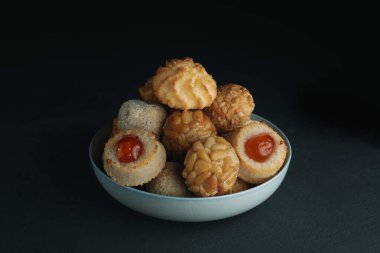 a blue ceramic bowl with an assortment of panellets, a typical confection of Catalonia, Spain, eaten traditionally on All Saints Day, placed on a dark green or dark gray table clipart