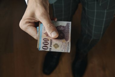 a businessman, wearing an elegant gray suit, offers a wad of hungarian forint notes to the observer, standing on a wooden floor indoors clipart