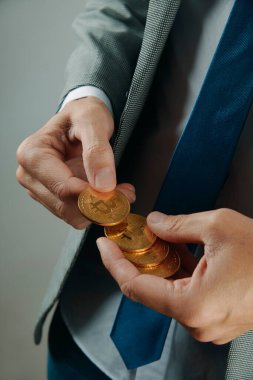 Gri takım elbiseli, beyaz gömlekli ve mavi kravatlı bir işadamı soluk gri bir arka planın önünde bitcoin atıyor.