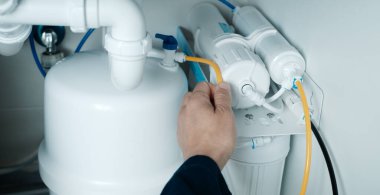 Mavi tulumlu beyaz bir adam mutfak lavabosunun altına ters osmoz sistemi kuruyor.