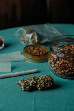 a cannabis bud on a table next to a spliff, a glass jar with some rolling tobacco, some sheets of rolling paper a used herb grinder and a plastic bag with some more cannabis buds clipart