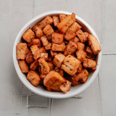 Beyaz bir seramik kasenin yüksek açılı görüntüsü. Kırsal beton yüzeyinde marine edilmiş tofu küpleri var.