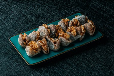 Yeşil dikdörtgen bir tabakta cevizlerle dolu kuru incirler. Masa seti üzerinde zarif, koyu yeşil bir masa örtüsü var.