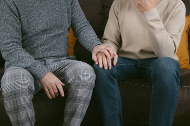 Gri pantolonlu ve gri kazaklı bir adam, yaralı bir adamın elini tutuyor, kot pantolon ve bej bir kazak giyiyor, ikisi de kahverengi bir kanepede oturuyor.