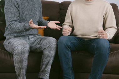 two men, wearing casual clothes, are arguing sitting on a brown sofa clipart