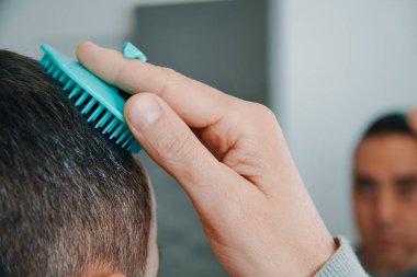 Bir adam saçlarını mavi yuvarlak bir saç fırçasıyla tarıyor. Banyoda aynanın önünde.