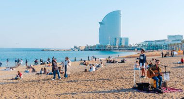 Barselona, İspanya - 28 Mart 2025: İspanya 'nın Barcelona kentindeki Sant Sebastia Plajı' nda bir bahar günü ve halk arasında Hotel Vela olarak bilinen W Barselona Oteli 'nde insanlar, arka planda ise yelkenli otel.
