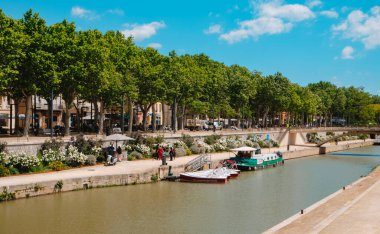 Narbonne, Fransa - 6 Mayıs 2025: Güneşli bir bahar gününde Fransa 'nın Narbonne şehrindeki Rue de la Republique' de ağaçlık gezinti alanı boyunca Kanal de la Robine manzarası