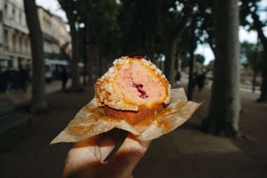 Elinde çilek reçeli dolu ısırılmış bir keki olan bir adamın yakın plan fotoğrafı. Yerel bir fırında yeni pişmiş, ağaçlarla kaplı bir güvertede duruyor.