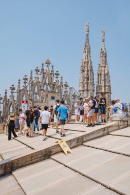 Milan, İtalya - 21 Haziran 2025: Turistler Milano Katedrali 'nin güneşli çatısında gotik kulelerin arasında yürürken eğik yüzey uyarısını görmezden geliyorlar.