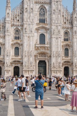 Milan, İtalya - 21 Haziran 2025: Meşgul bir meydanda duran mavi gömlekli bir adam, açık yaz gökyüzünün altındaki ikonik gotik cepheyi fotoğraflamak için telefonunu kaldırırken Milan Katedraline bakıyor.