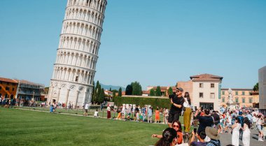 Pisa, İtalya - 26 Haziran 2025: Bir adam bir eliyle Pisa Kulesi 'ni engellemek için tırabzanda duruyor, kalabalık bir meydanda açık mavi gökyüzü altında poz veren düzinelerce turistin arasına katılıyor.