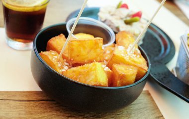 Bir kase İspanyol patatesi bravas, baharatlı soslu kızarmış patates, arka planda bir içki ve bir tabak ensaladilla rusa ile birlikte bir restoran masasında kaba tuz ile servis edilir.