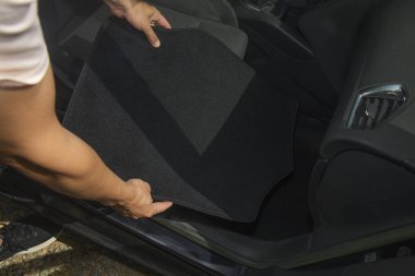 Ön kapıdan bakıldığında, bir adam yeni siyah bir paspası arabanın iç tarafındaki halının üzerine indiriyor, yolcu tarafını tamamen kaplaması için dikkatlice hizalıyor.