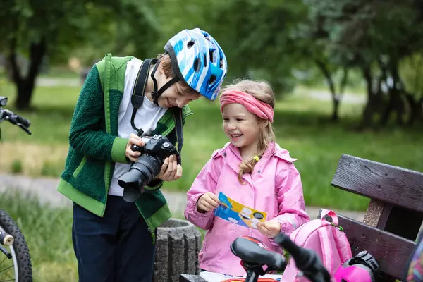 若い男の子と女の子は カメラのディスプレイと笑顔を見ています 兄は妹に連れて行かれた写真を見せている 公園で自転車に乗って話したり笑ったりする子供たち ロイヤリティフリーのストック画像