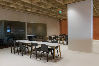 Bir sürü sandalye ve masa olan bir oda. Sandalyeler siyah ve masalar beyaz. Oda boş ve çok temiz ve düzenli görünüyor.