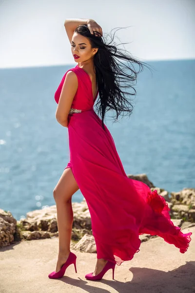 海岸の赤い長いドレスを着た美しい黒髪の女の子は夏の日を楽しんでいます — ストック写真
