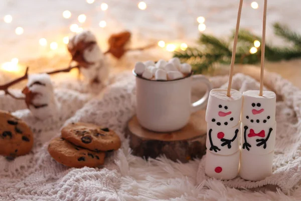 Cozy winter or Christmas background with funny marshmallow snowmen, cup of hot chocolate and cookies on white warm blanket. Cozy atmosphere.