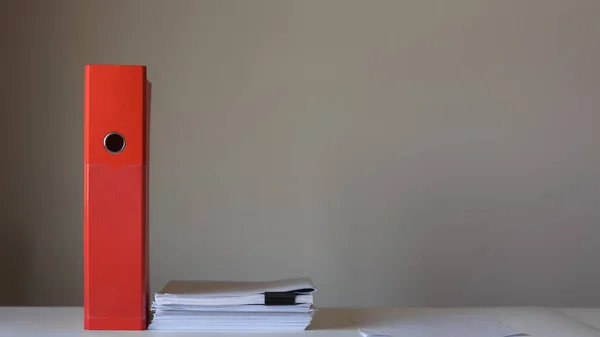 Red folder and stack of papers on empty wall background.