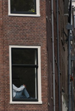 Person in white sweater and blue jeans sitting on window sill of a brick building on Brouwersgracht, Amsterdam clipart