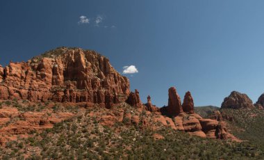 Sedona, Arizona 'daki yüksek kızıl kaya kuleleri ve uçurumların manzarası. Engebeli kumtaşı oluşumları, bir tutam bulutla berrak bir mavi gökyüzünün altında Amerikan Sou 'sunun saf güzelliğini yansıtan çöl bitkisinin üzerine çarpıcı bir şekilde yükselir.