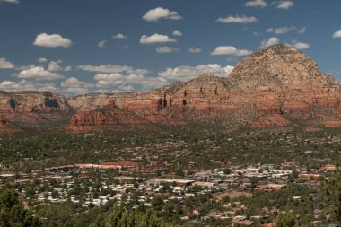 Sedona, Arizona 'nın panoramik manzarası, kasabanın ön planda olması ve arka plandaki dramatik kırmızı kaya oluşumlarının küçük beyaz bulutlarla benekli canlı mavi gökyüzünün altında yükselmesi..