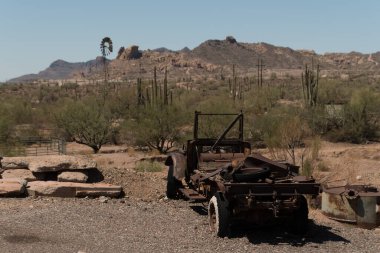 Eski paslı bir araç iskeleti Goldfield Hayalet Kasabası yakınlarındaki çölde terk edilmiş bir şekilde duruyor, arkasında dağlar ve kaktüsler var. Bu görüntü, Amerika 'nın güneybatısındaki engebeli tarih ve madencilik kültürünü vurguluyor..