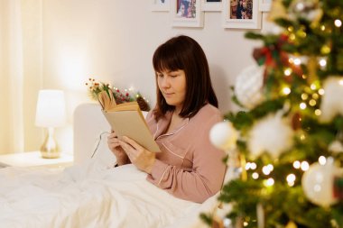 Süslü yatak odasında Noel ağacıyla kitap okuyan genç bir kadının portresi.