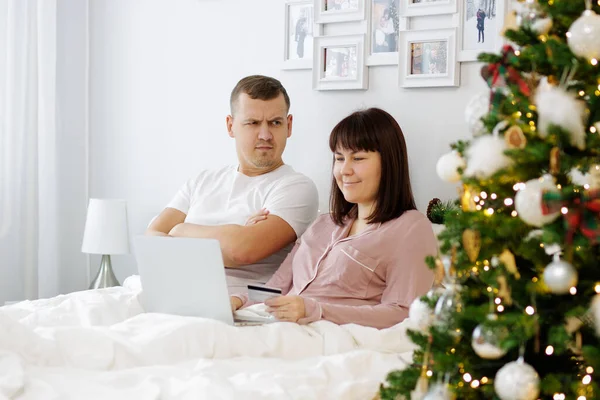 オンラインショッピング 幸せな女性はベッドの上に座って オンラインで何かを購入し 彼女の驚きの夫はクリスマスツリーで装飾された寝室で — ストック写真