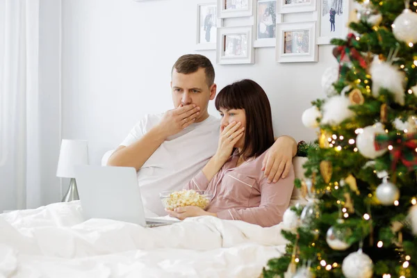 驚くべきカップルの肖像画ベッドに座ってクリスマスツリーで装飾されたベッドルームでラップトップで映画を見て — ストック写真