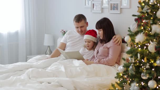 爱情和圣诞节的概念 年轻的父母在装饰过的圣诞树旁的卧室里给小女儿读书 — 图库视频影像