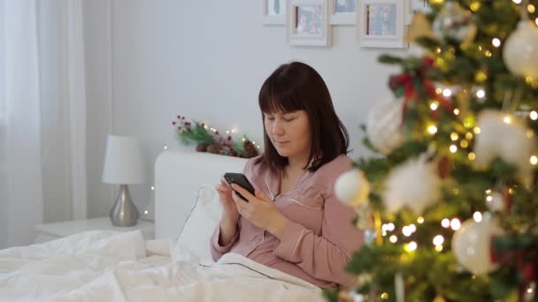 家和圣诞节的概念 穿着睡衣的年轻女人坐在床上 在装饰过的圣诞树旁使用智能手机 — 图库视频影像