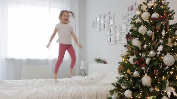 童年和圣诞节的概念 可爱的小女孩在装饰过的圣诞树旁跳上了床 — 图库视频影像