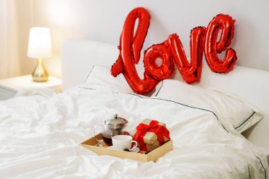 Sevgililer Günü ve romantik konsept. Bir fincan çay, çaydanlık, hediye kutusu ve yatak odasındaki aşk sözcüğünün olduğu kırmızı balon içeren ahşap tepsi.