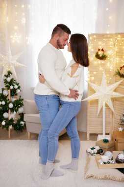 Genç evli bir çift Noel süslemeli odada birbirine sarılıp burunlarına dokunuyor.