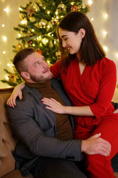 赤いスーツの女が男の足に座ってクリスマスツリーの近くに彼を見て — ストック写真