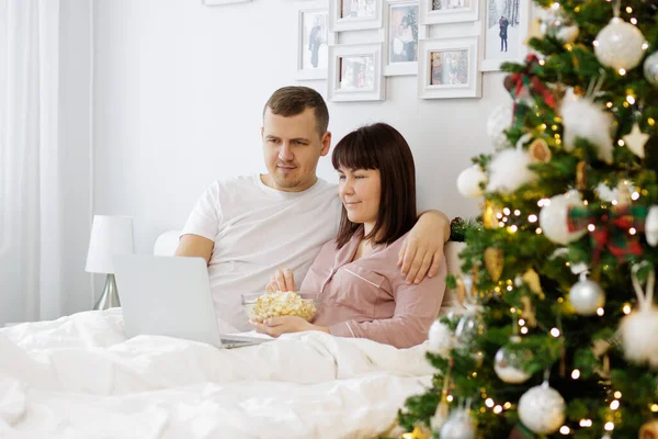 幸せなカップルの肖像画ベッドの上に座って クリスマスツリーと装飾されたベッドルームでラップトップで映画を見て — ストック写真